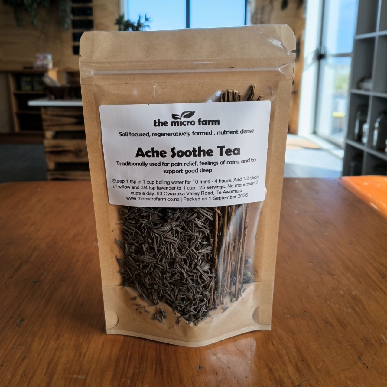 A stand-up pouch of 'Ache Soothe Tea' by 'the micro farm' sitting on a wooden table in a home setting. The pouch is made of a natural kraft paper material, with a white label showing the product details, and tea is visible through a clear window at the bottom.