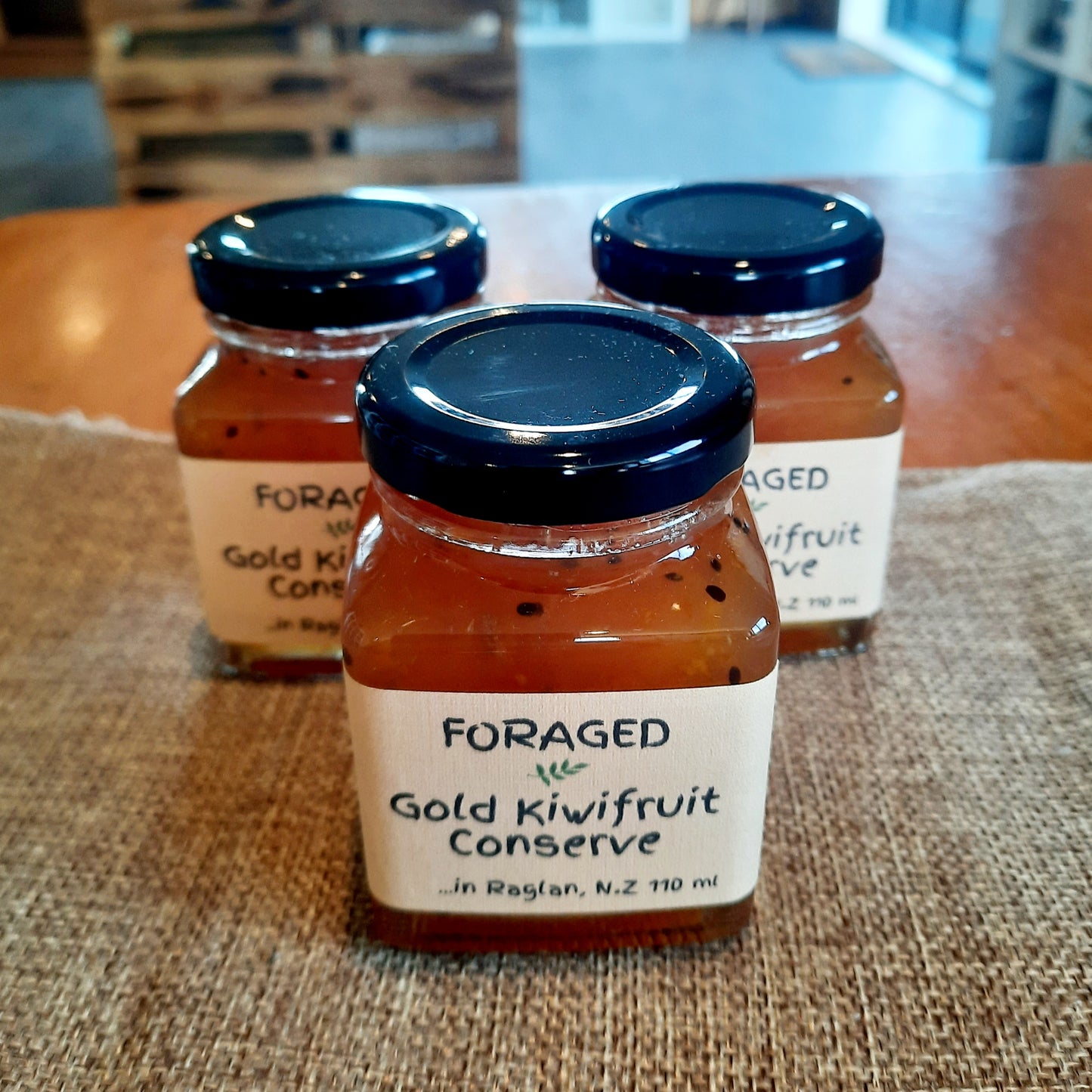 Three glass jars of Foraged Gold Kiwifruit Conserve with dark blue lids, arranged on a burlap surface on a wooden table.