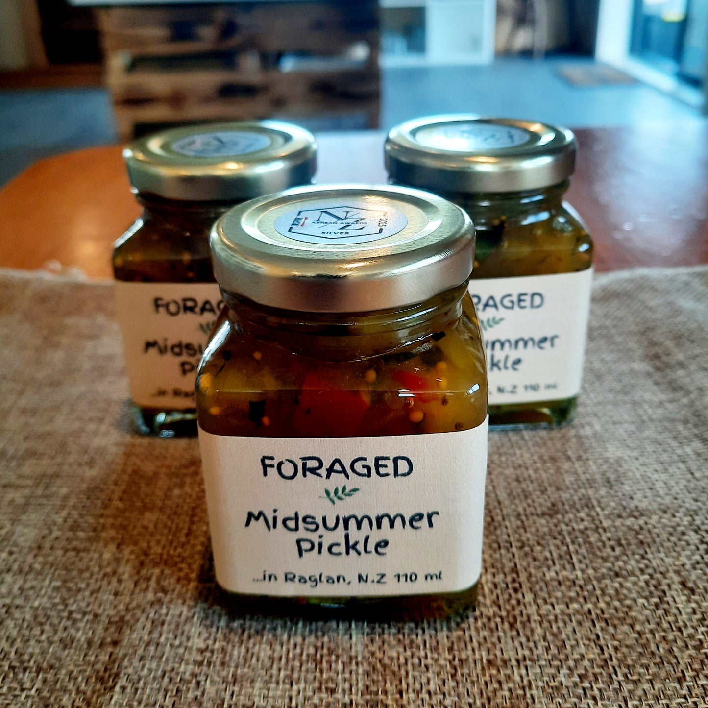 Three glass jars of Foraged in Raglan Midsummer Pickle arranged on a burlap surface, with the front jar in sharp focus and the back jars softly blurred in a real indoor setting.