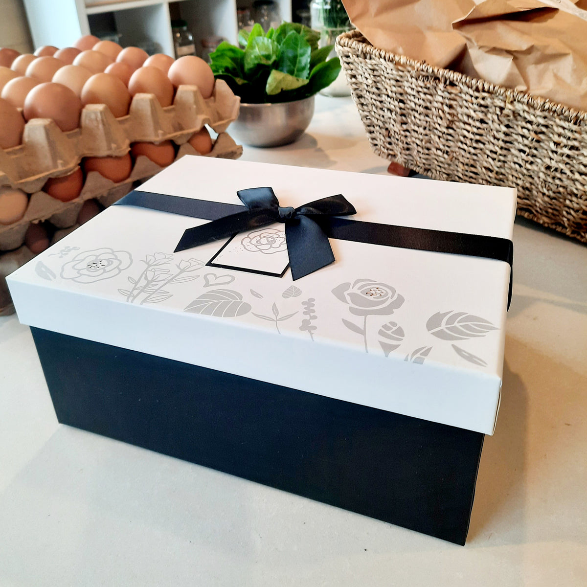 A rectangular gift box with a white lid decorated with gray floral line art and a black ribbon tied in a bow, set on a kitchen counter with eggs and a basket in the background.