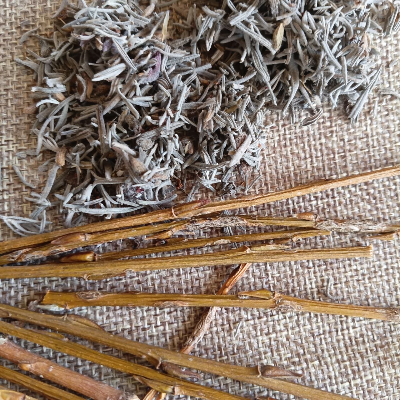 Dried botanical ingredients, including a pile of finely chopped pale gray-green lavender leaves and several long tan-brown willow stems, arranged on a woven beige burlap surface.