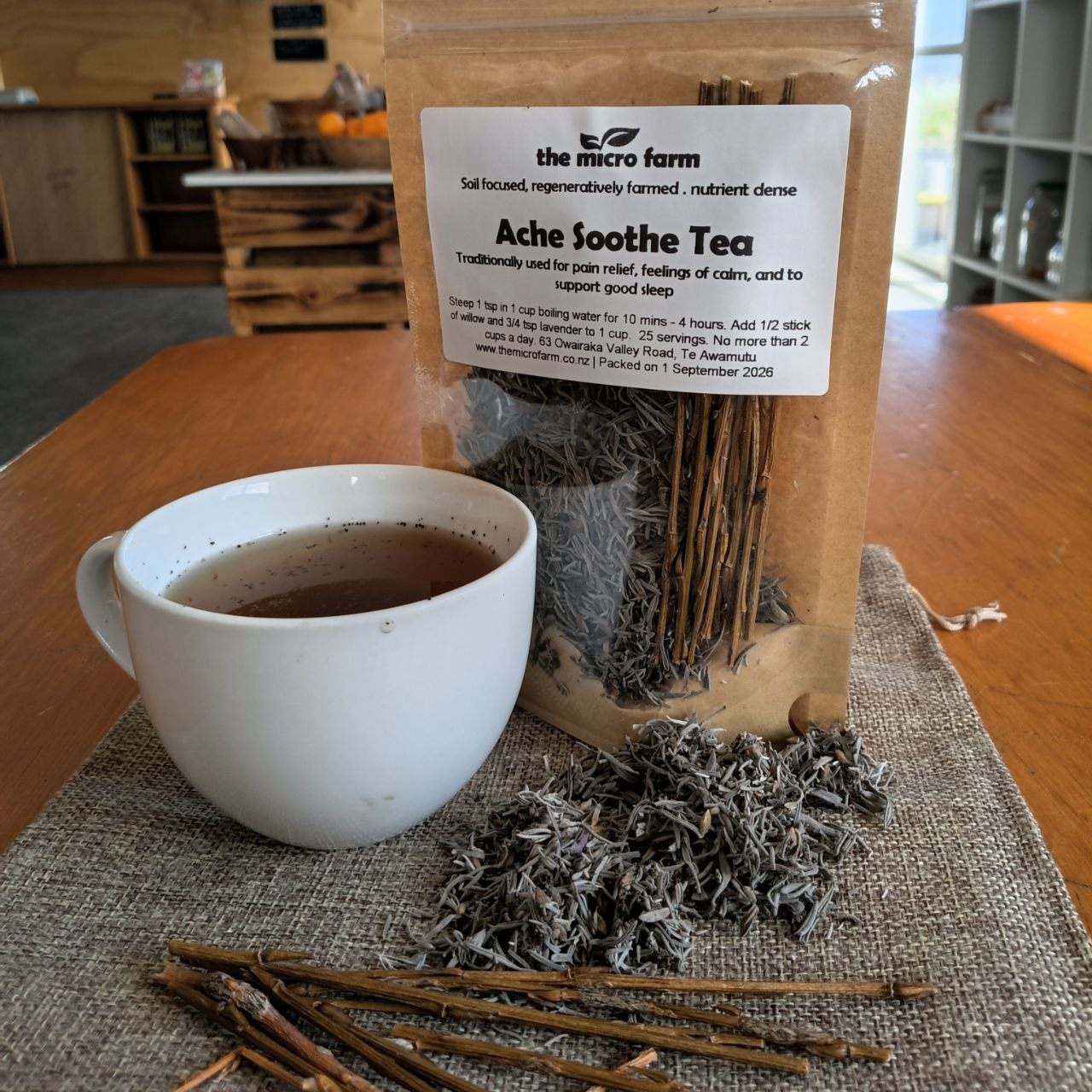 A clear stand-up pouch labeled 'the micro farm Ache Soothe Tea' sits on a wooden table beside a white cup of tea and loose dried tea leaves on a burlap runner.