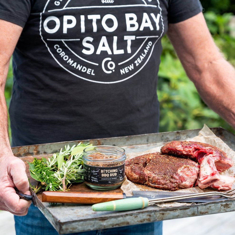 Person holding a tray with raw meat, herbs, and a jar of Opito Bay Salt Co. product.
