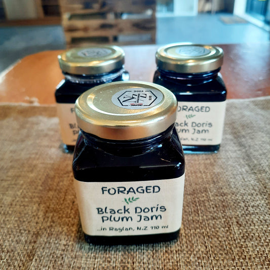 Three glass jars of Foraged Black Doris Plum Jam arranged on a burlap surface with a wooden table in the background.