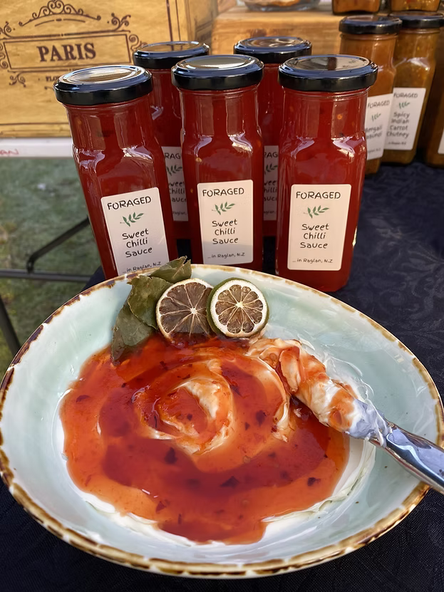 A bowl of Foraged Sweet Chili Sauce with cream, lime halves, and a bay leaf, with several glass jars of the same sauce lined up behind it on a dark tablecloth.