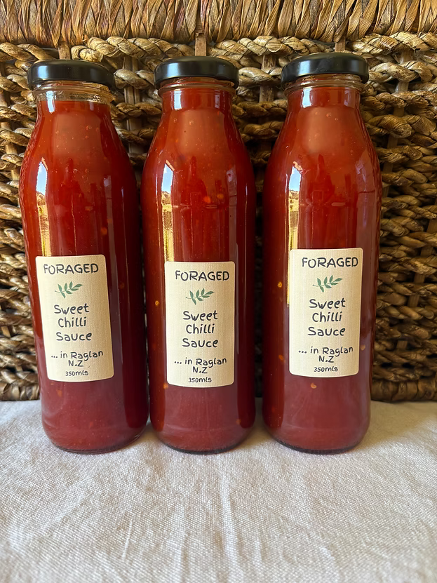 Three glass bottles of Foraged Sweet Chilli Sauce lined up against a woven wicker basket background, resting on a light beige fabric surface.