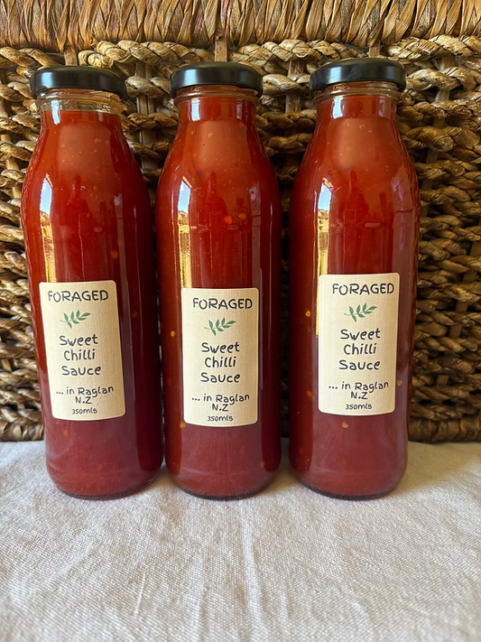 Three glass bottles of Foraged Sweet Chilli Sauce lined up against a woven wicker basket background, resting on a light beige fabric surface.