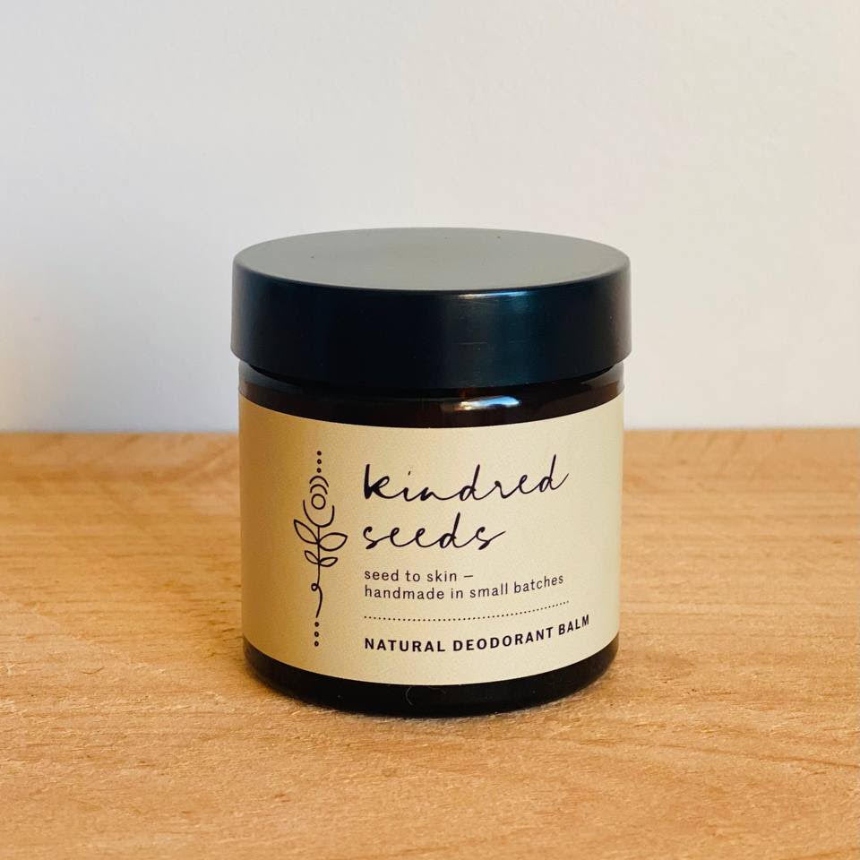A jar of Kindred Seeds Natural Deodorant Balm sits on a light wood surface against a plain white wall. The product features a black lid, a dark amber glass jar, and a beige label with black text.