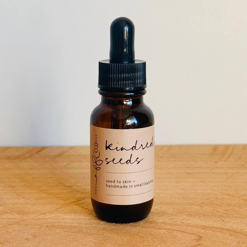An amber glass dropper bottle with a black cap and a cream label reading 'Kindred Seeds' and 'seed to skin - handmade in small batches,' standing on a light wood surface against a plain off-white wall.