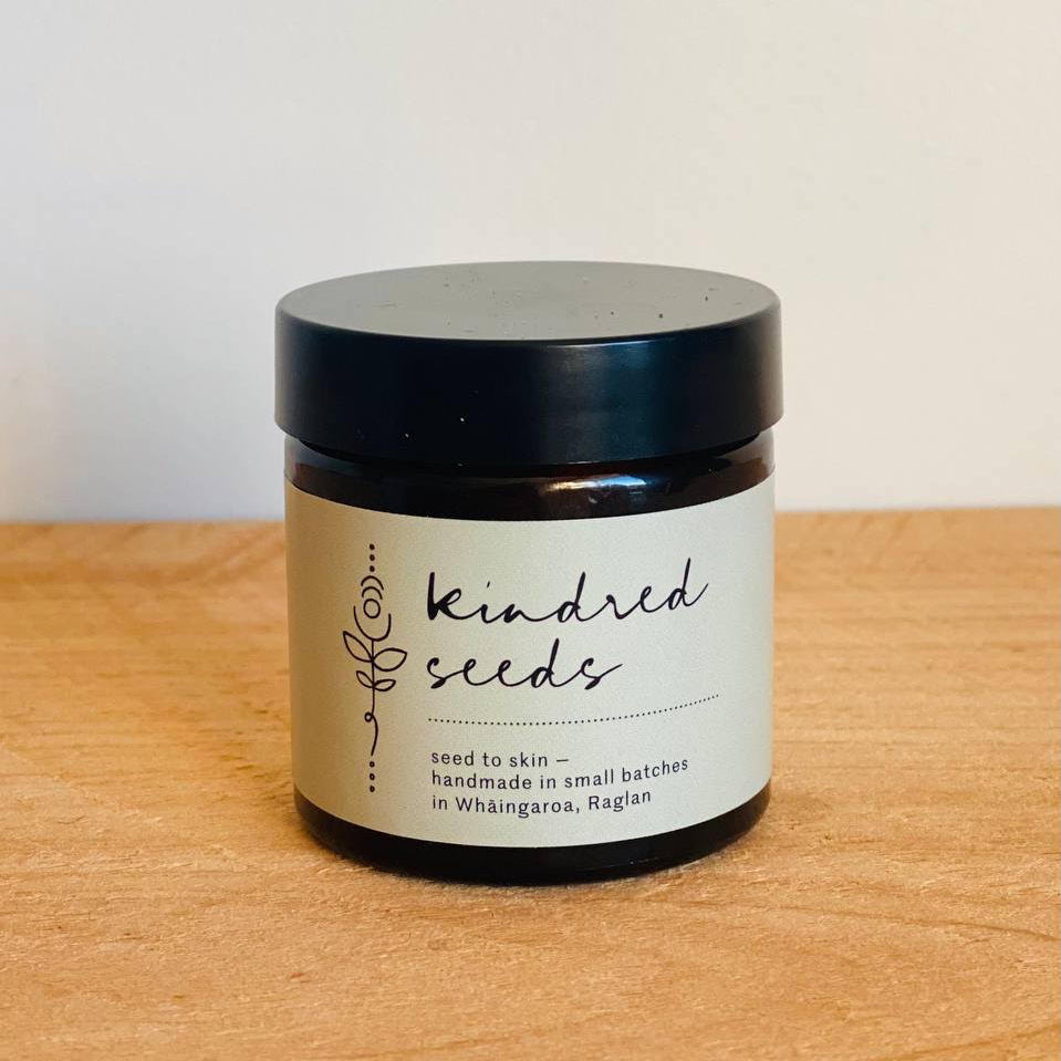 A small dark amber jar with a black lid and a cream label reading 'kindred seeds' sits on a light wood surface against a plain off-white wall.