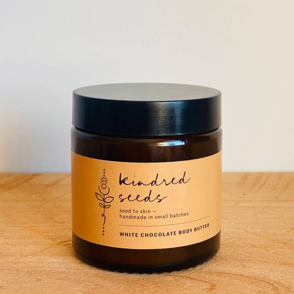 A jar of Kindred Seeds White Chocolate Body Butter sitting on a light wood surface against a plain off-white wall. The product is in a dark amber glass jar with a black lid and a tan paper label featuring black text.