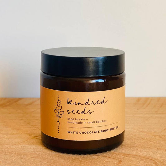 A jar of Kindred Seeds White Chocolate Body Butter sitting on a light wood surface against a plain off-white wall. The product is in a dark amber glass jar with a black lid and a tan paper label featuring black text.