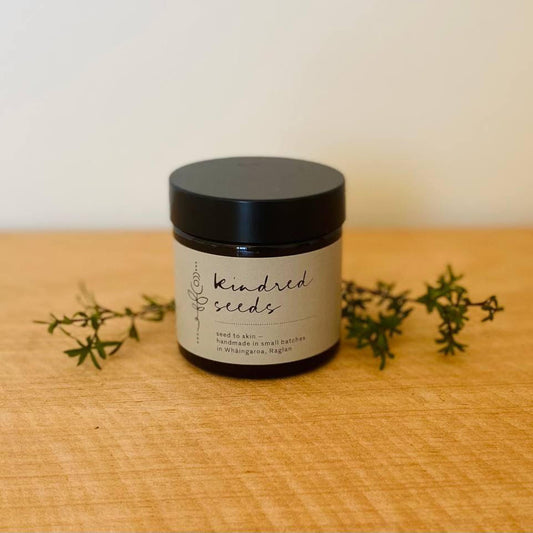 A small black jar labeled 'Keenred Seeds' sits on a light wood surface with small green sprigs placed around it against a plain light wall.