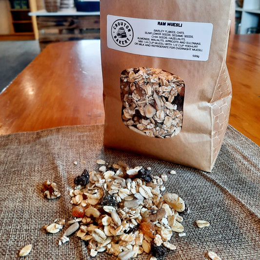 A 500g paper bag of Crouton Bakery Raw Muesli with a clear window showing the granola inside, placed on a wooden table with some of the granola spilled in front on a burlap runner.
